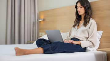 Een persoon zittend op een bed met een laptop, in een kamer met een houten hoofdeinde en een gordijn.