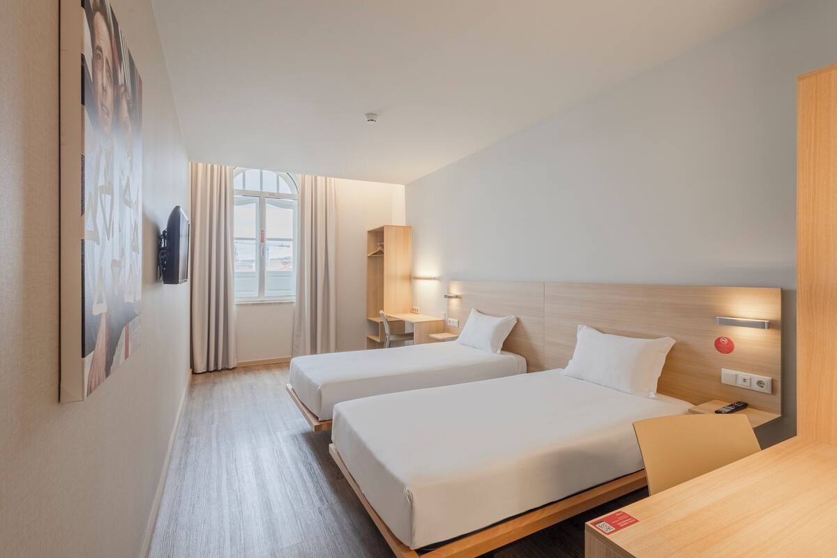 A hotel room with two beds, a desk, a chair, a nightstand, and a window with curtains.