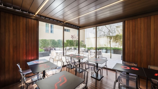 An outdoor dining area with wooden paneling, glass partitions, and tables with red accents. - Moov Hotel Porto Centro (Porto)