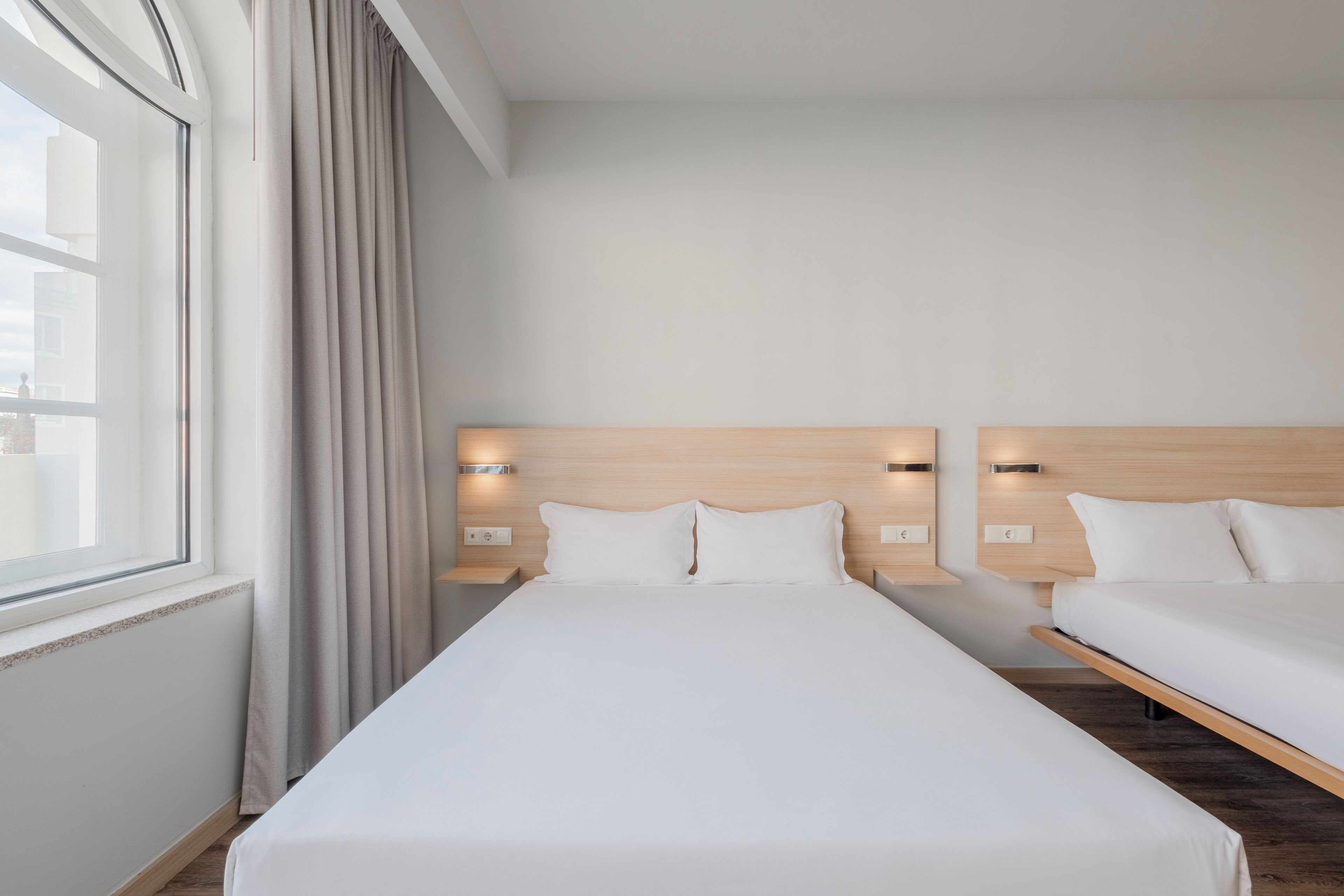 A hotel room with two beds, a large window with curtains, and wall-mounted lighting.