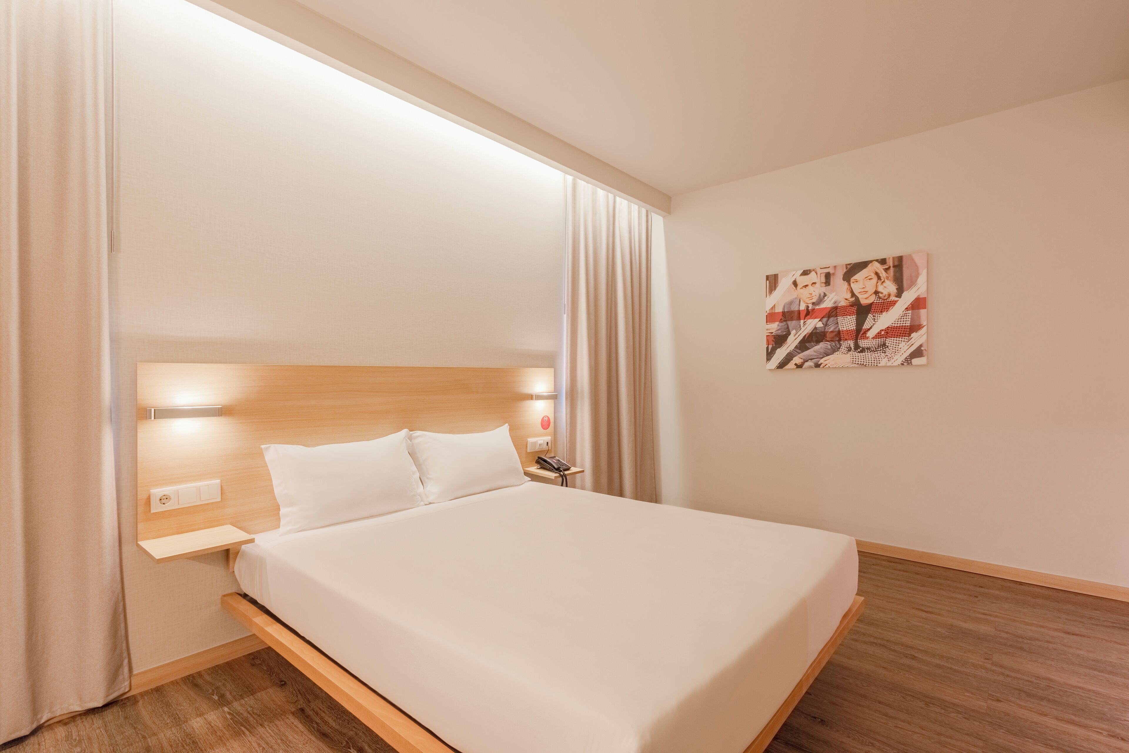 A hotel room with a wooden headboard, a white bed, a nightstand with a phone, and a painting on the wall.