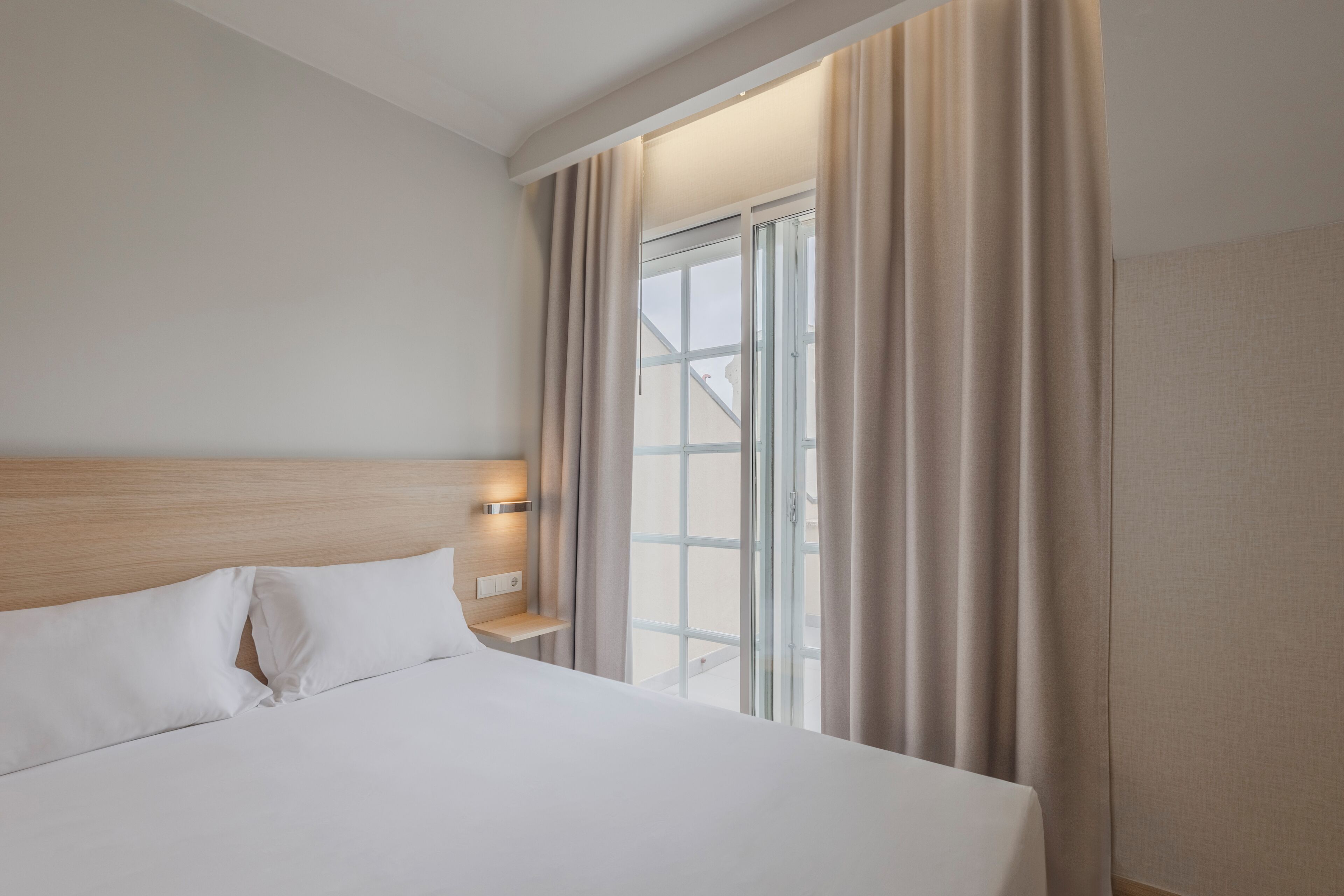A hotel room with a large bed, a nightstand, a lamp, and a view of a building through a window with curtains.