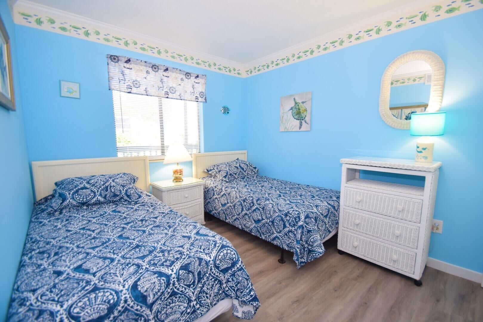Une chambre avec deux lits, une table de chevet avec une lampe, une commode, une fenêtre avec des stores et un miroir fixé au mur.