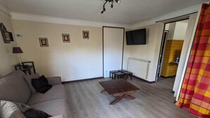 A living room with a sofa, a coffee table, a TV mounted on the wall, and a door leading to another room. - Charming T1 of 30 m² on the ground floor of villa (Toulon)