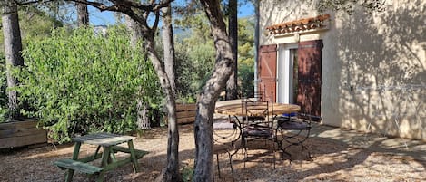 Un espacio al aire libre con mesa y sillas de madera, rodeado de árboles y suelo de grava.