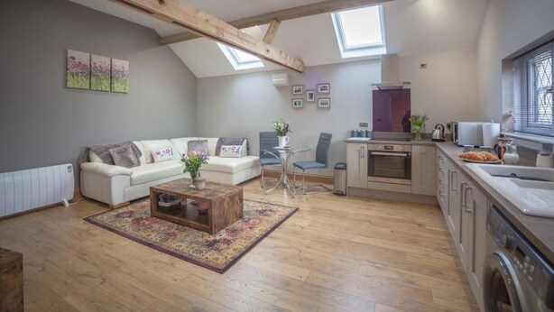 A spacious loft with a high ceiling, exposed wooden beams, a cozy living area with a sofa and coffee table, a small dining table, and a compact kitchen with a window overlooking the city. - Little Oak (Hereford)