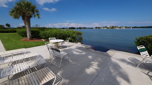 Um pátio externo com cadeiras e mesas de metal branco, uma palmeira e vista para um lago com edifícios ao longe.