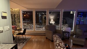 Una sala de estar con un sofá, dos sillones, una mesa de centro y una mesa de comedor. Hay ventanas amplias con luces decorativas.