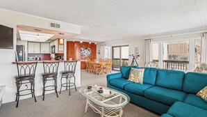 Ein modernes Wohnzimmer mit einem türkisblauen Sofa, einem Glastisch und einer Küchenzeile mit Bar.