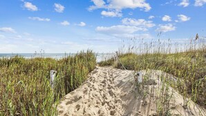 Ein Weg durch Sand am Strand, gesäumt von hohem Gras, mit blauem Himmel.