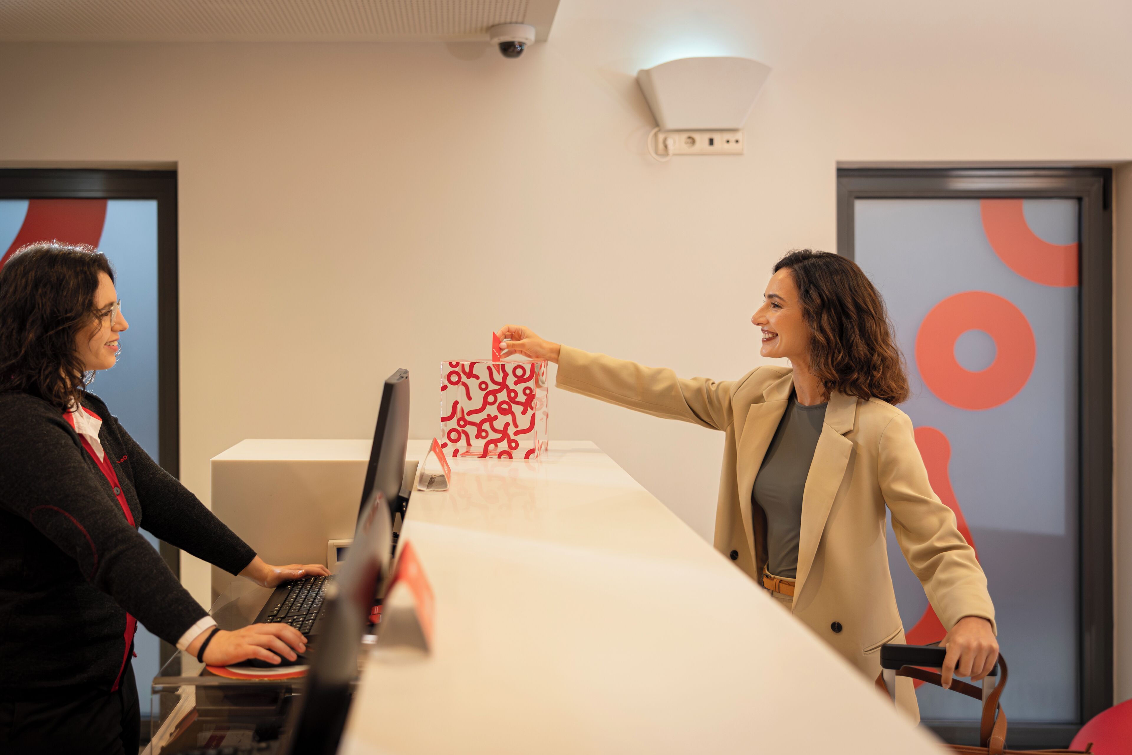 Two people at a reception desk in an office setting with a computer and a framed artwork on the wall.