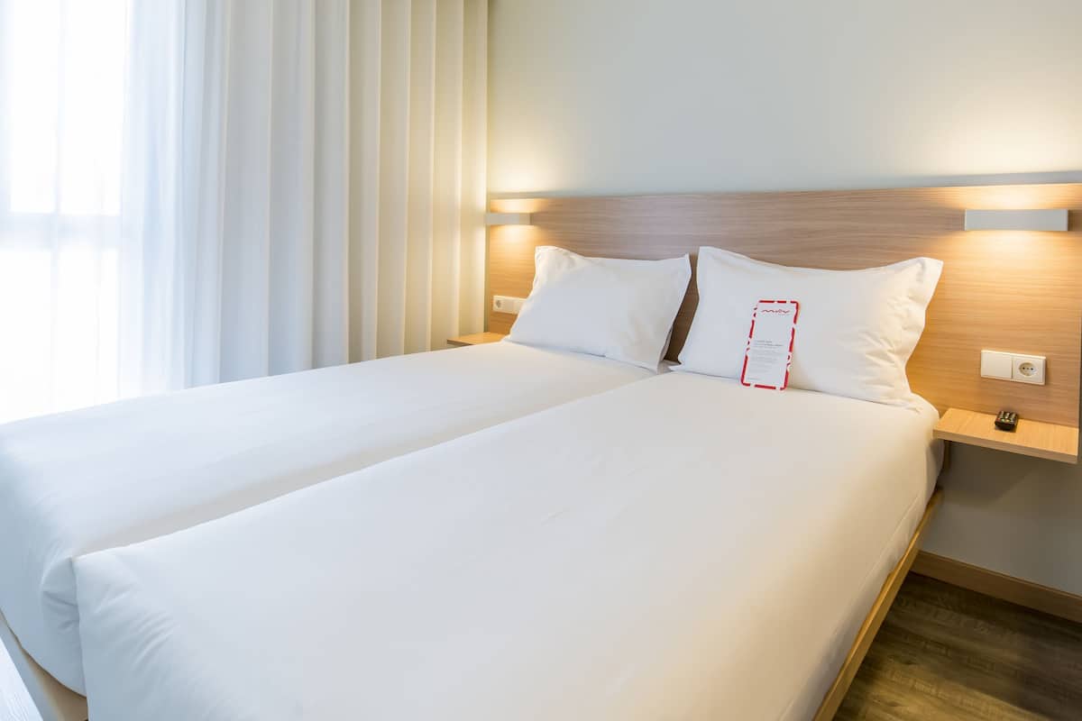 A hotel room with a large bed, a nightstand, a wall-mounted light, and a window with curtains.