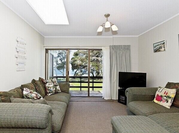 A living room with two green sofas, a flat-screen TV, and a large window with a view of trees and a lawn. - Beachside at Snells - Snells Beach Apartment (Snells Beach)