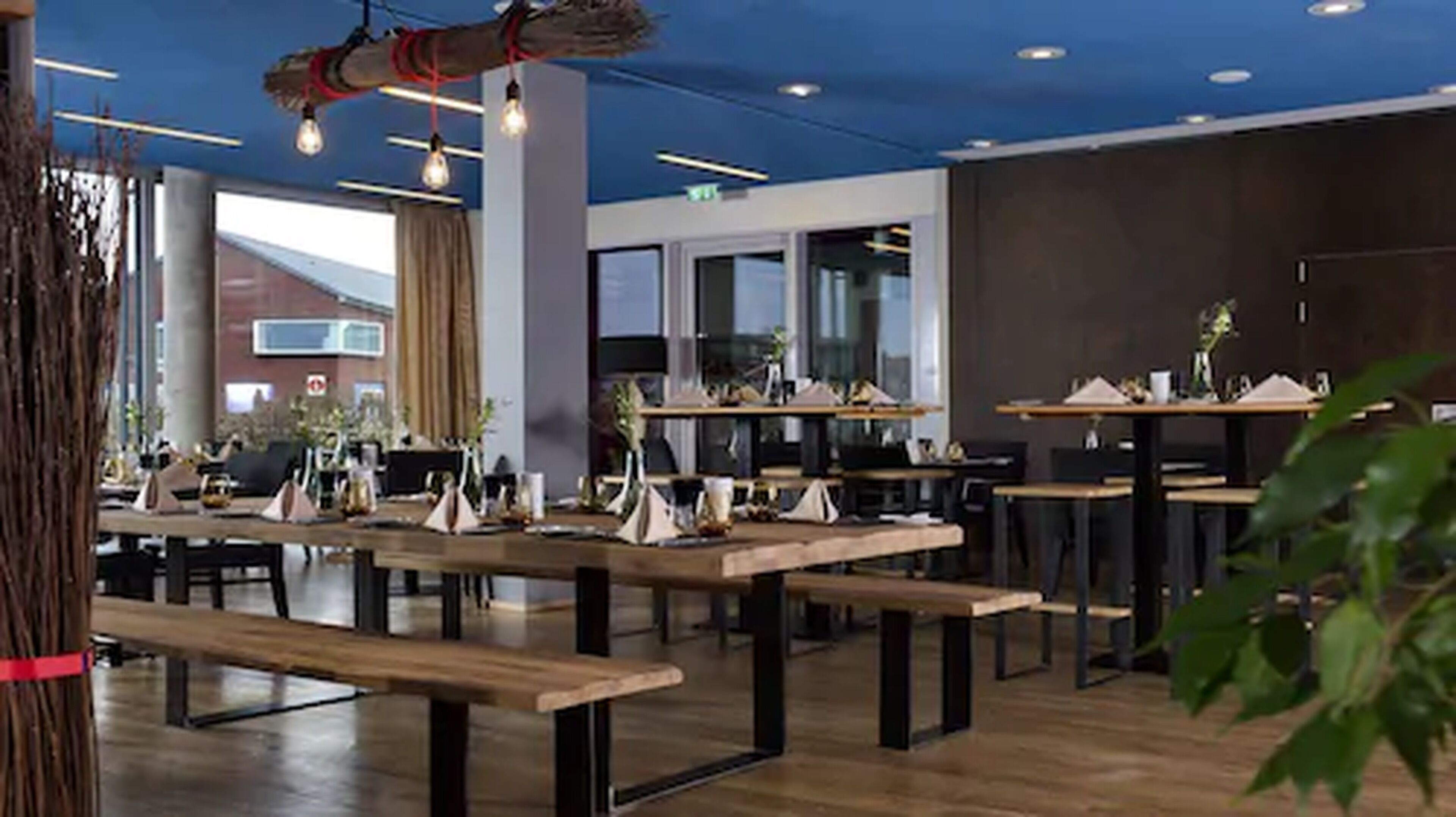 Een moderne eetruimte met houten tafels en banken, grote ramen en hangende lampen.