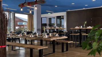 Een moderne eetruimte met houten tafels en banken, grote ramen en hangende lampen.