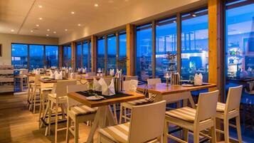 A modern restaurant with high ceilings and large windows offering city views. Tables are neatly arranged with white chairs.