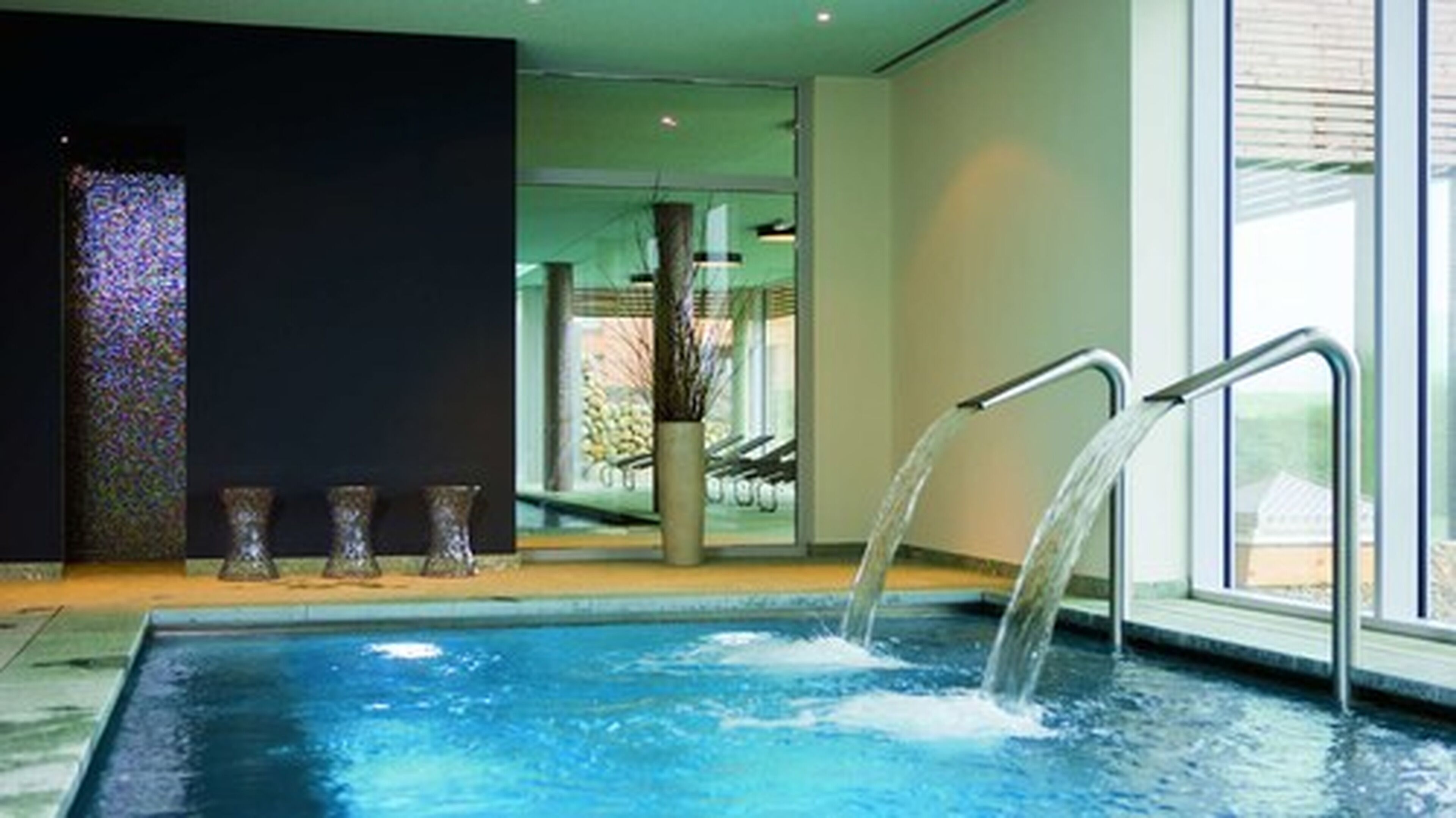 Une piscine agrémentée d’un élément décoratif aquatique, un mur foncé orné d’un panneau décoratif et une porte vitrée donnant sur un espace extérieur.