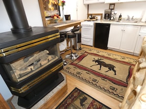 Una cocina con estufa de leña, armarios blancos, cafetera y un tapete con diseños de animales.