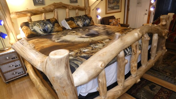 Un cadre de lit en bois rustique, surmonté d’une couverture à motif de loup, avec une lampe et un miroir.
