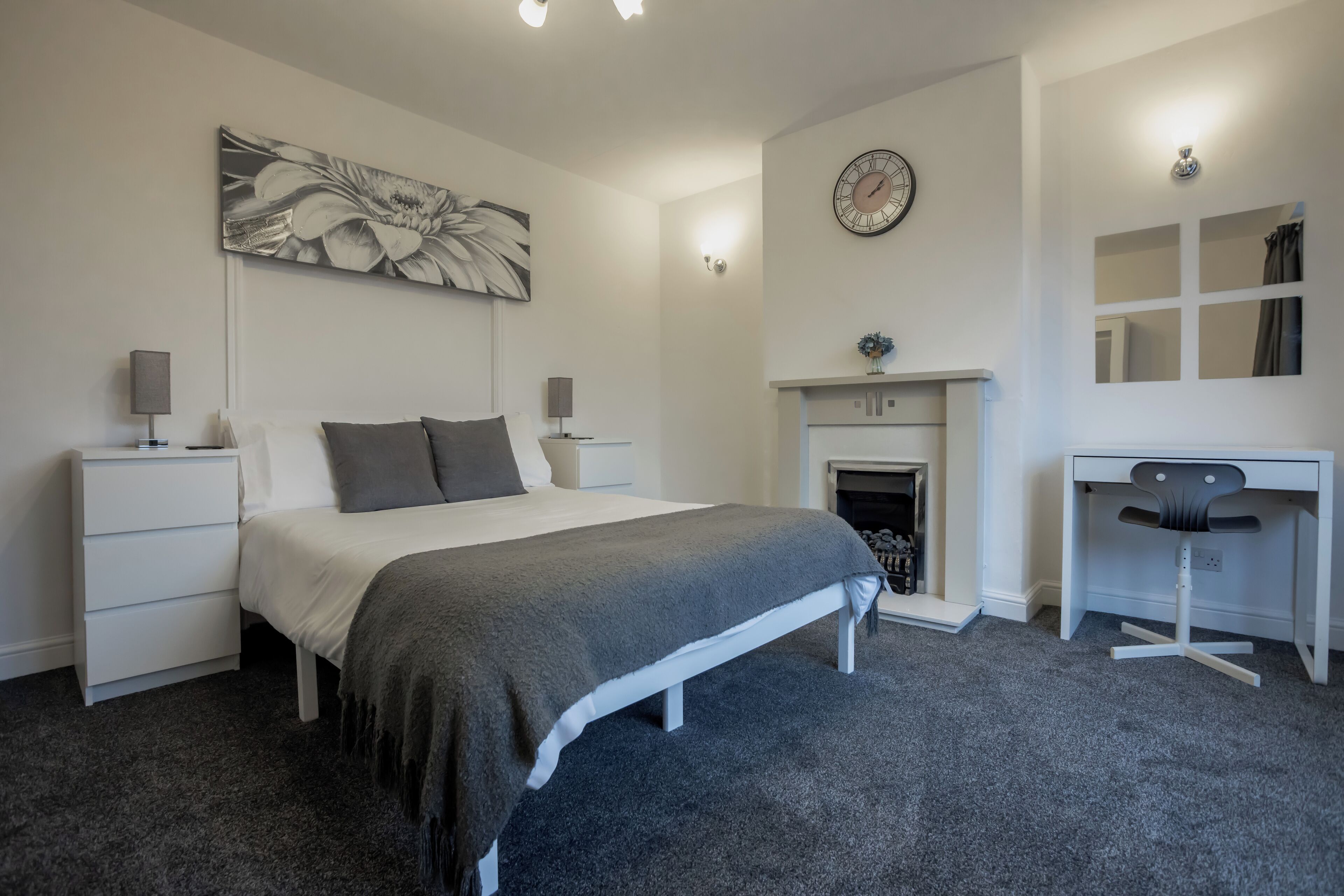 A bedroom with a bed, bedside tables, a desk, a chair, a fireplace, and a wall clock.