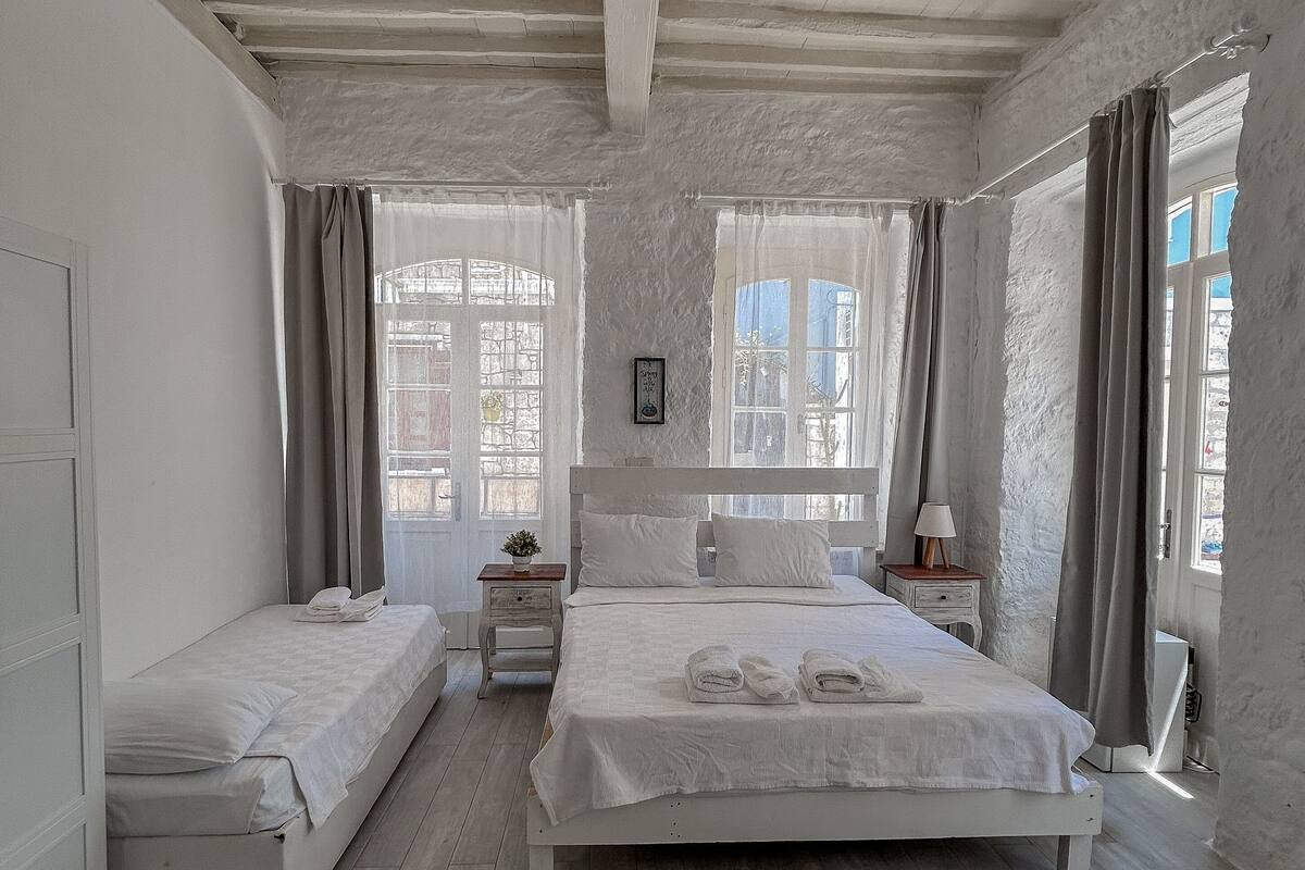 A bedroom with two beds, a wooden ceiling, and large windows with curtains.