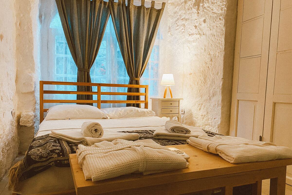 A bed with a wooden headboard, a bedside table with a lamp, and a window with curtains.