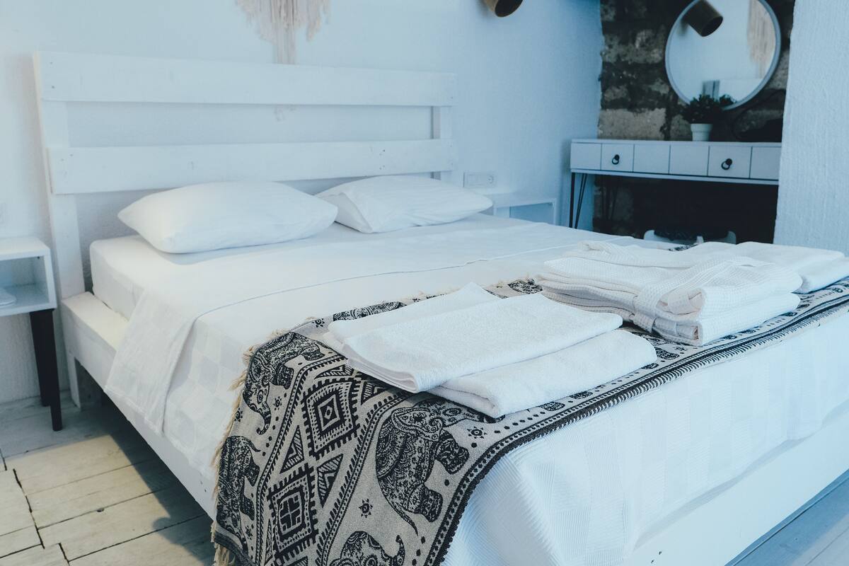 A neatly made bed with white linens and a patterned throw blanket, a bedside table, a dresser with a mirror, and a wall-mounted macrame piece.