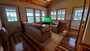 Ein gemütliches Wohnzimmer mit braunem Ledersofa, einem Fernseher und großen Fenstern mit Blick ins Freie.