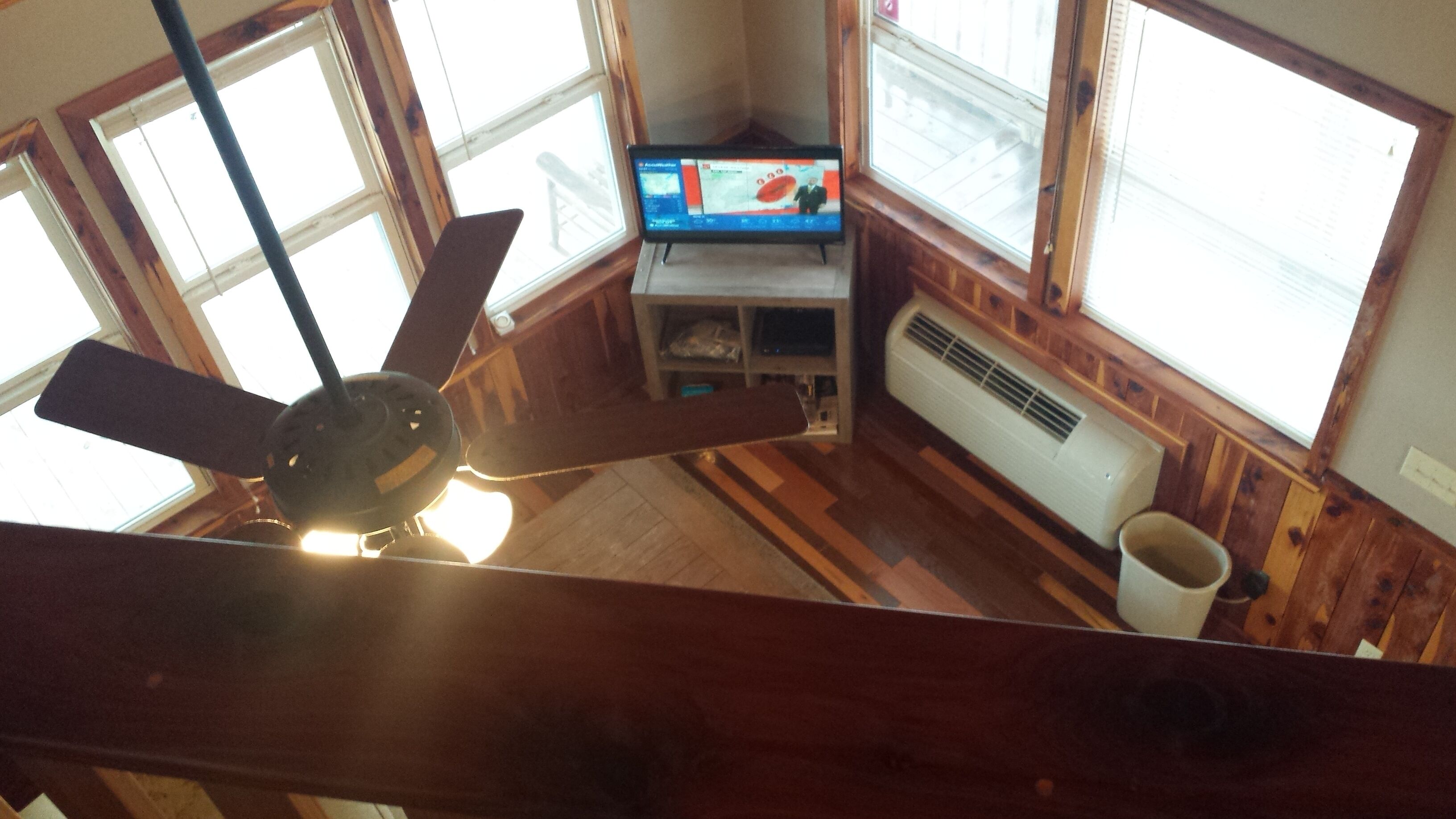 A room with a ceiling fan, a television on a stand, a white radiator, and large windows.