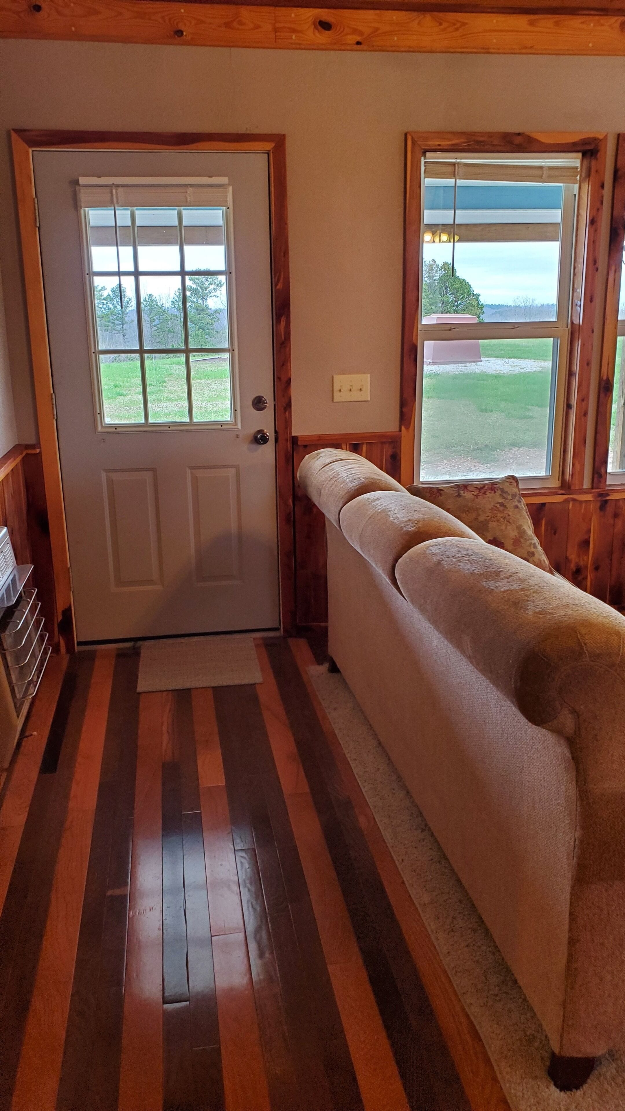 A room with a wooden floor, a door with a view of the outdoors, a window with a view of the outdoors, and a bench with a cushion.