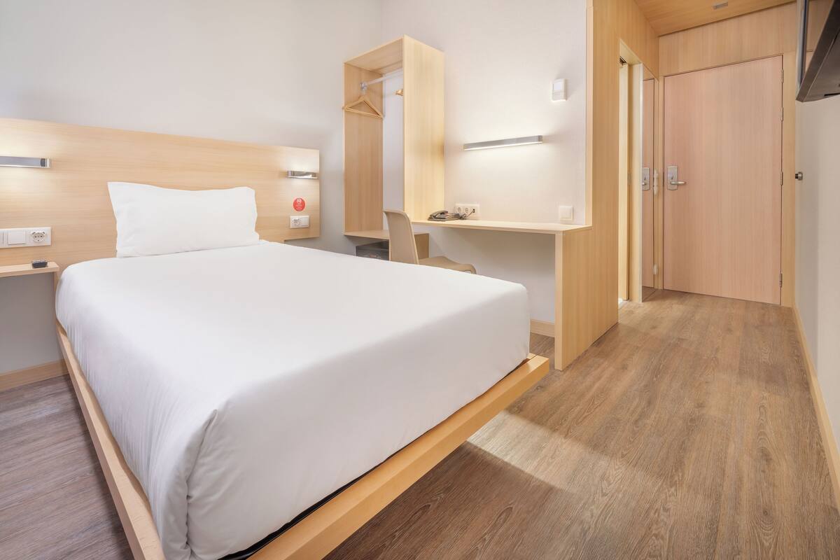 A modern hotel room with a large bed, wooden headboard, bedside table, and a built-in desk with a chair.