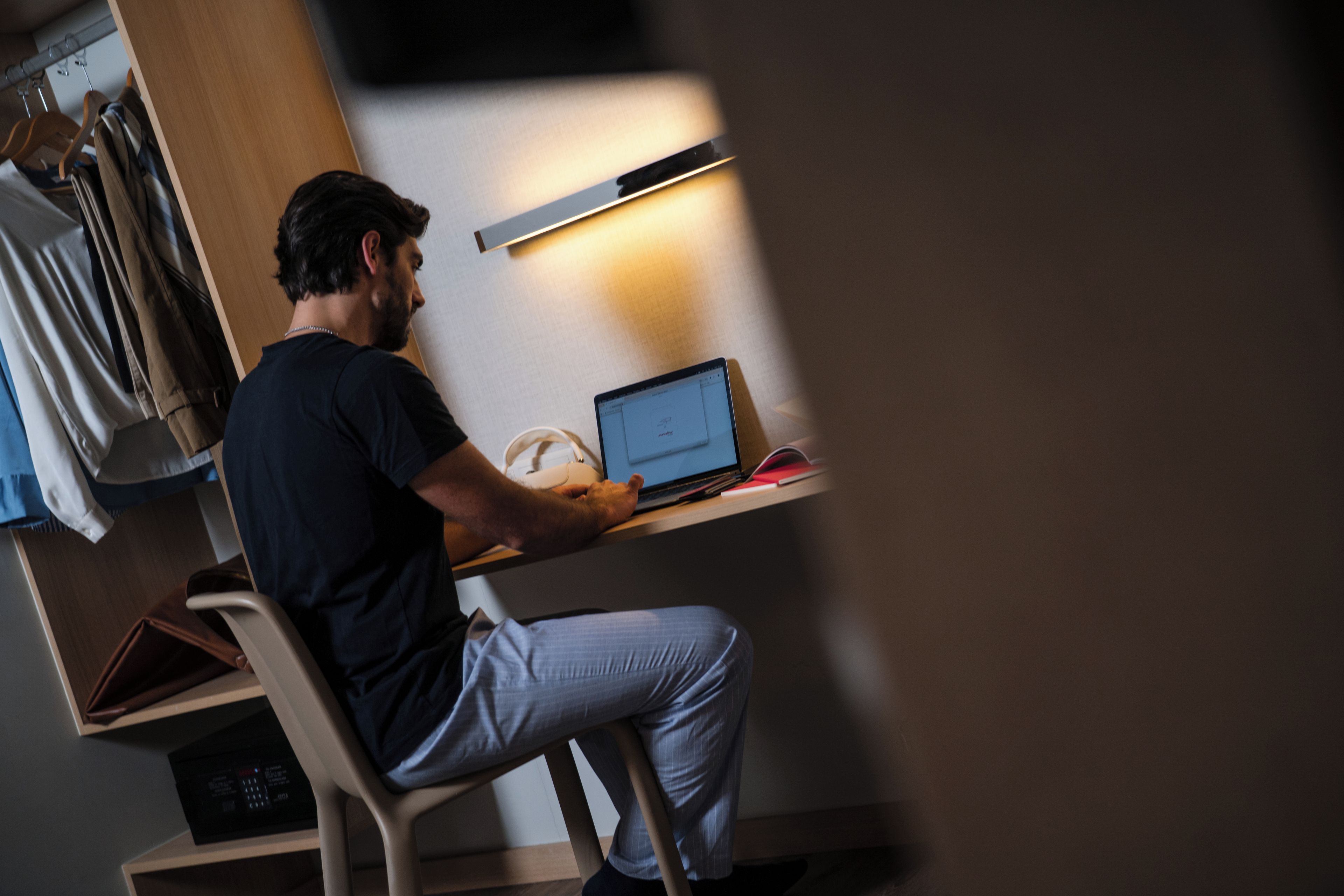 A person sitting at a desk with a laptop, a white cup, and a red book. There is a wooden chair and a shelf with a black object on it.