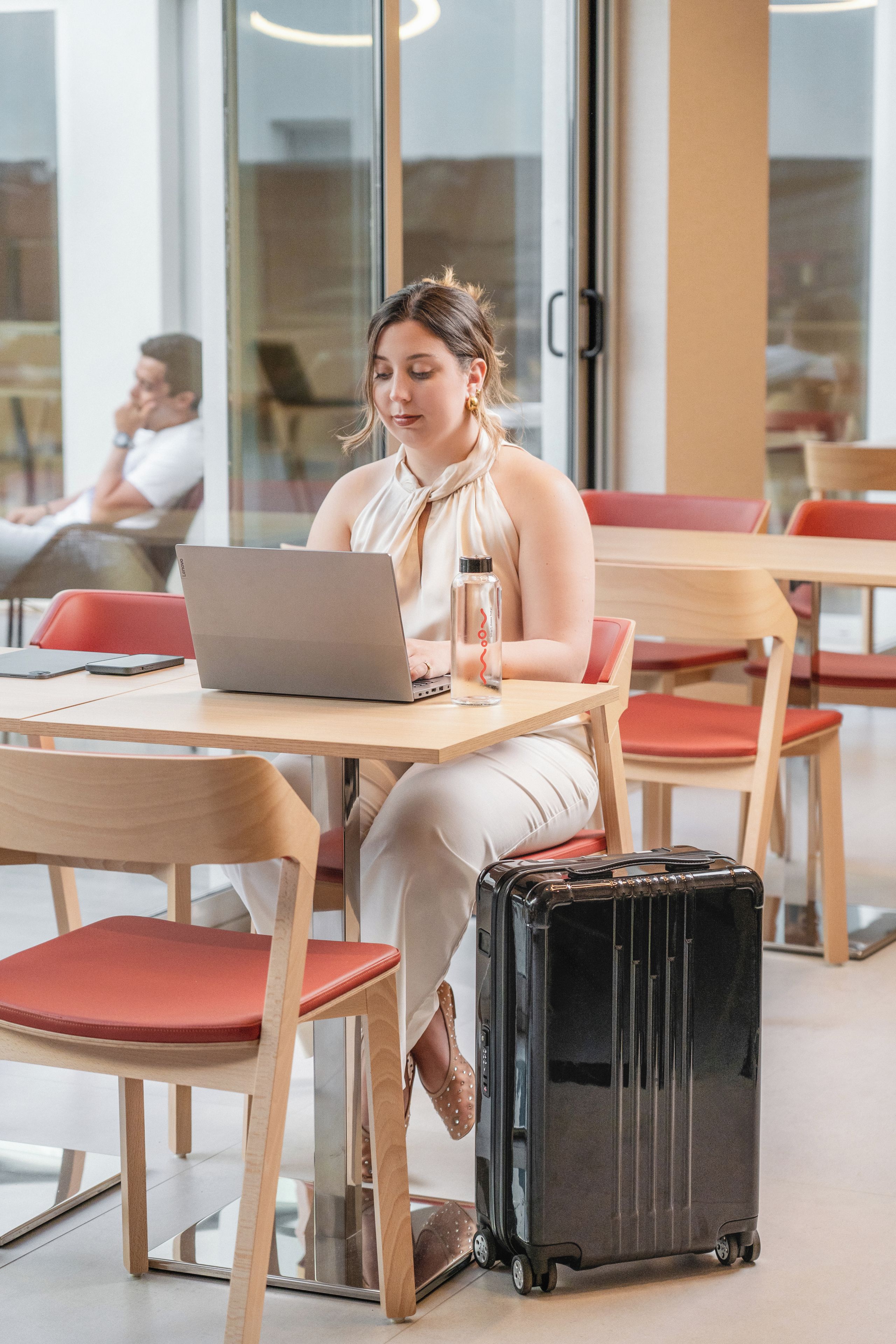 A person seated at a table with a laptop, a bottle, and a suitcase.
