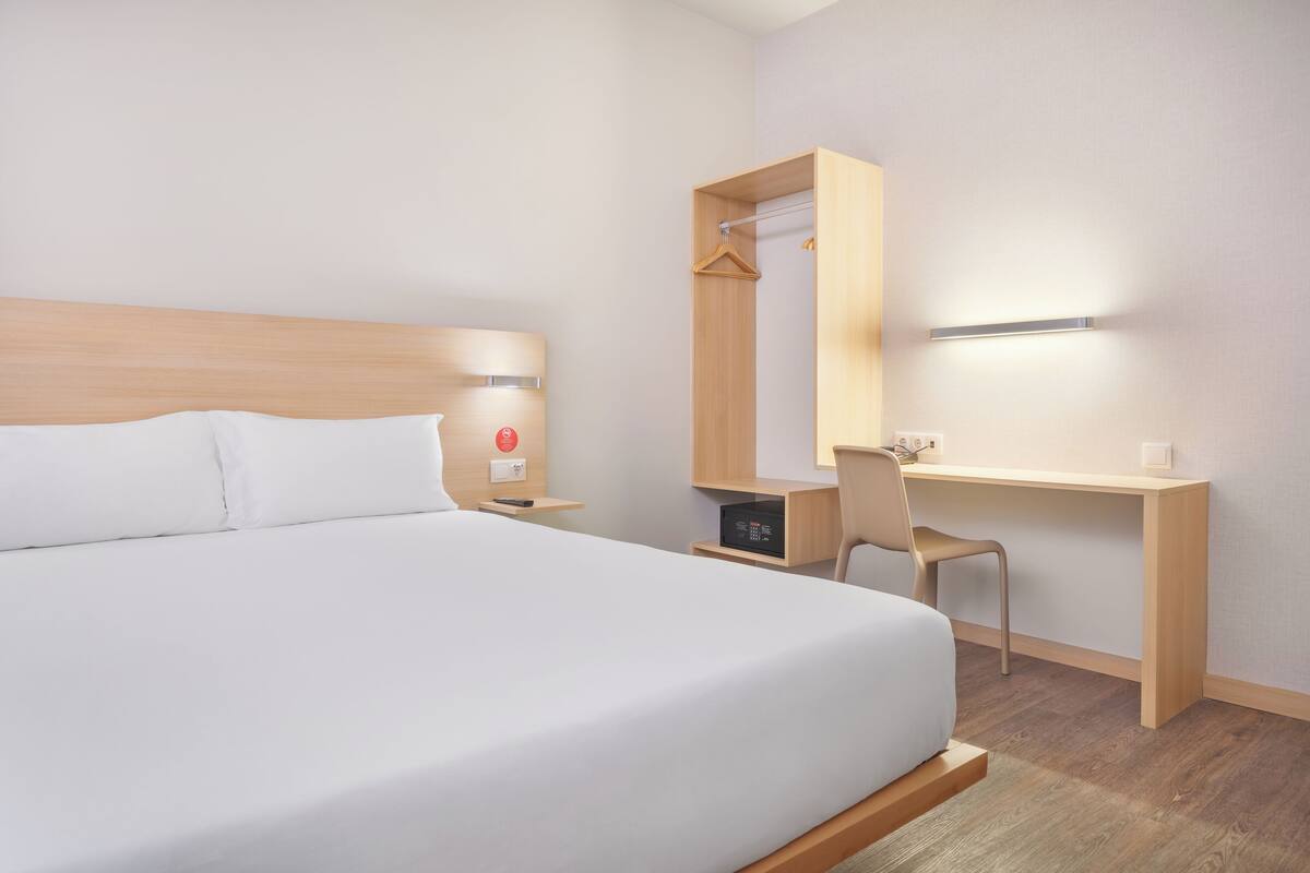 A hotel room with a bed, a desk, a chair, a mirror, and a bedside table with a lamp.