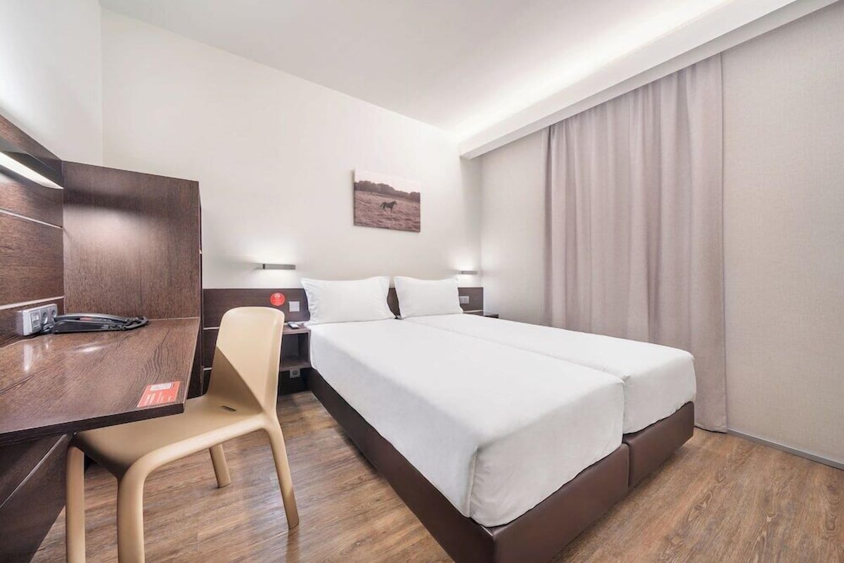 Ein Hotelzimmer mit Bett, Schreibtisch, Stuhl, Nachttisch, Lampe und einem Gemälde an der Wand.
