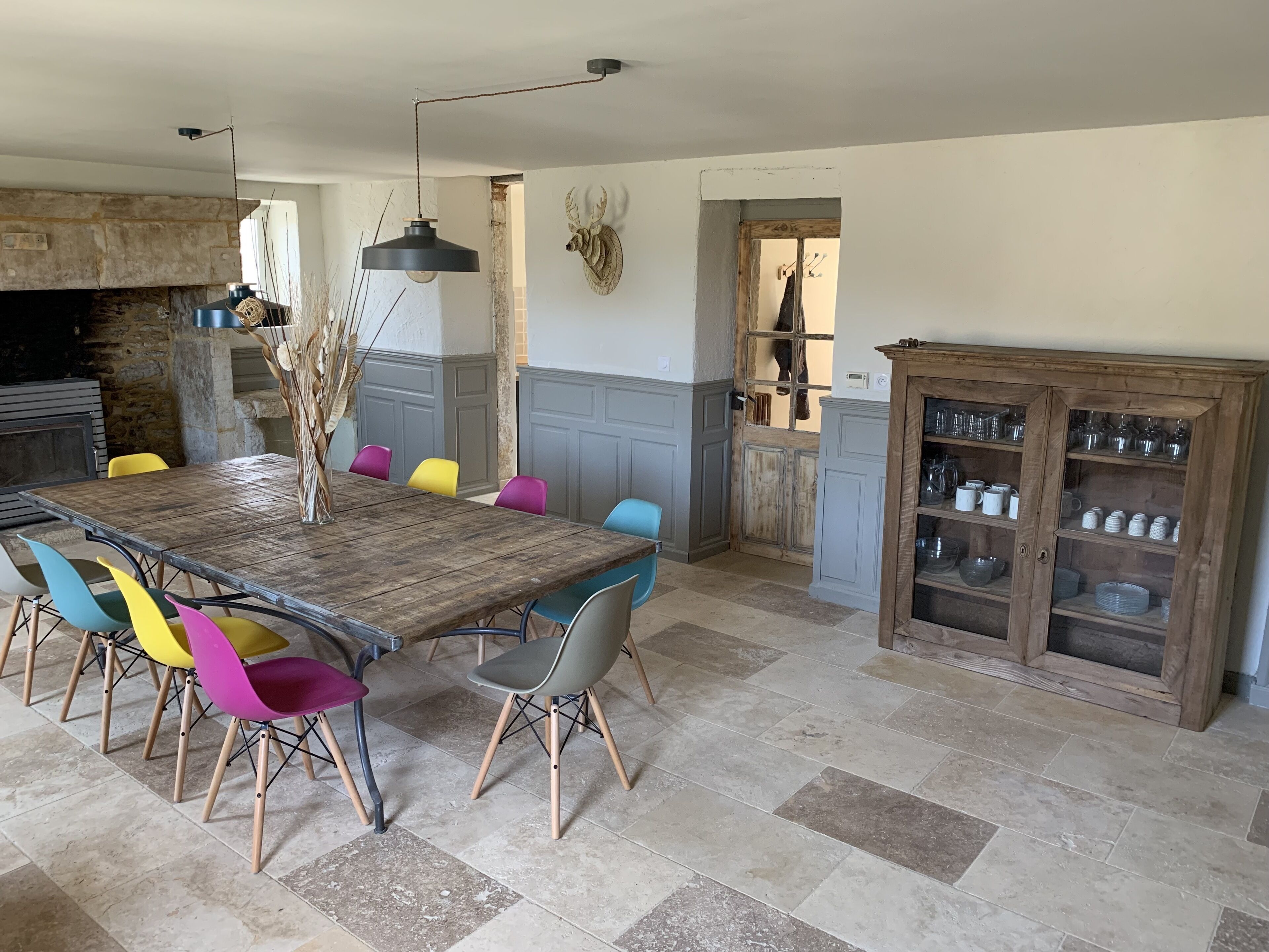 A dining area with a wooden table and colorful chairs, a stone fireplace, and a cabinet with glassware.