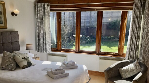 A bedroom with a large bed, a chair, a nightstand, and a window with curtains. - Whortleberry Studio - delightful bright airy ensuite B&B (Porlock)