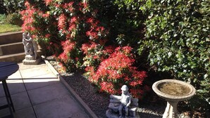 A garden with a statue of a man and woman on a bench, a birdbath, and red flowering bushes. - Whortleberry Studio - delightful bright airy ensuite B&B (Porlock)