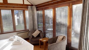A hotel room with a bed, two armchairs, a small table, and large windows with blinds. - Whortleberry Studio - delightful bright airy ensuite B&B (Porlock)