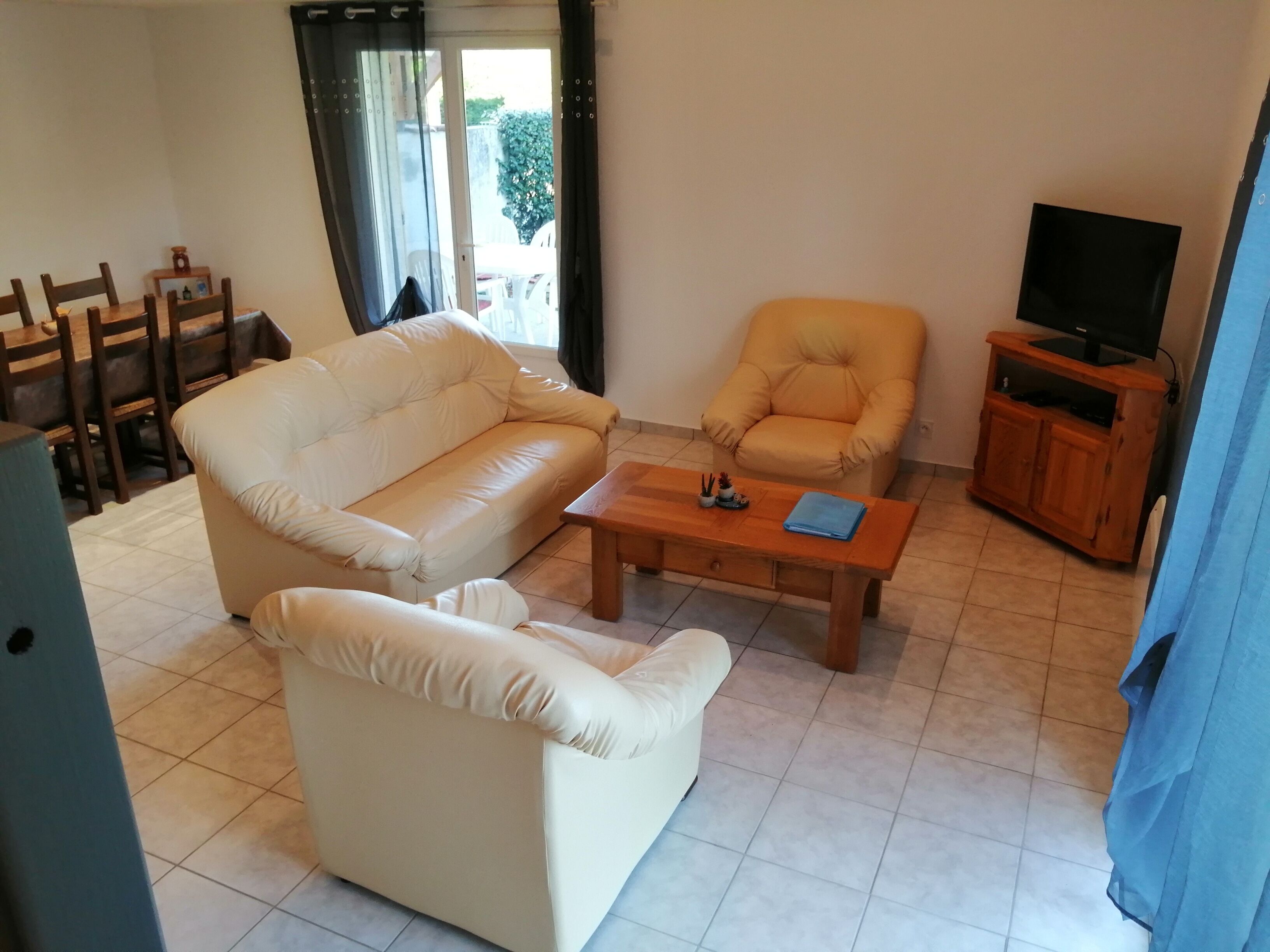Una sala de estar con un sofá color beige, un sillón a juego, una mesa de centro de madera, un televisor sobre un mueble de madera, y un comedor con sillas y una mesa.