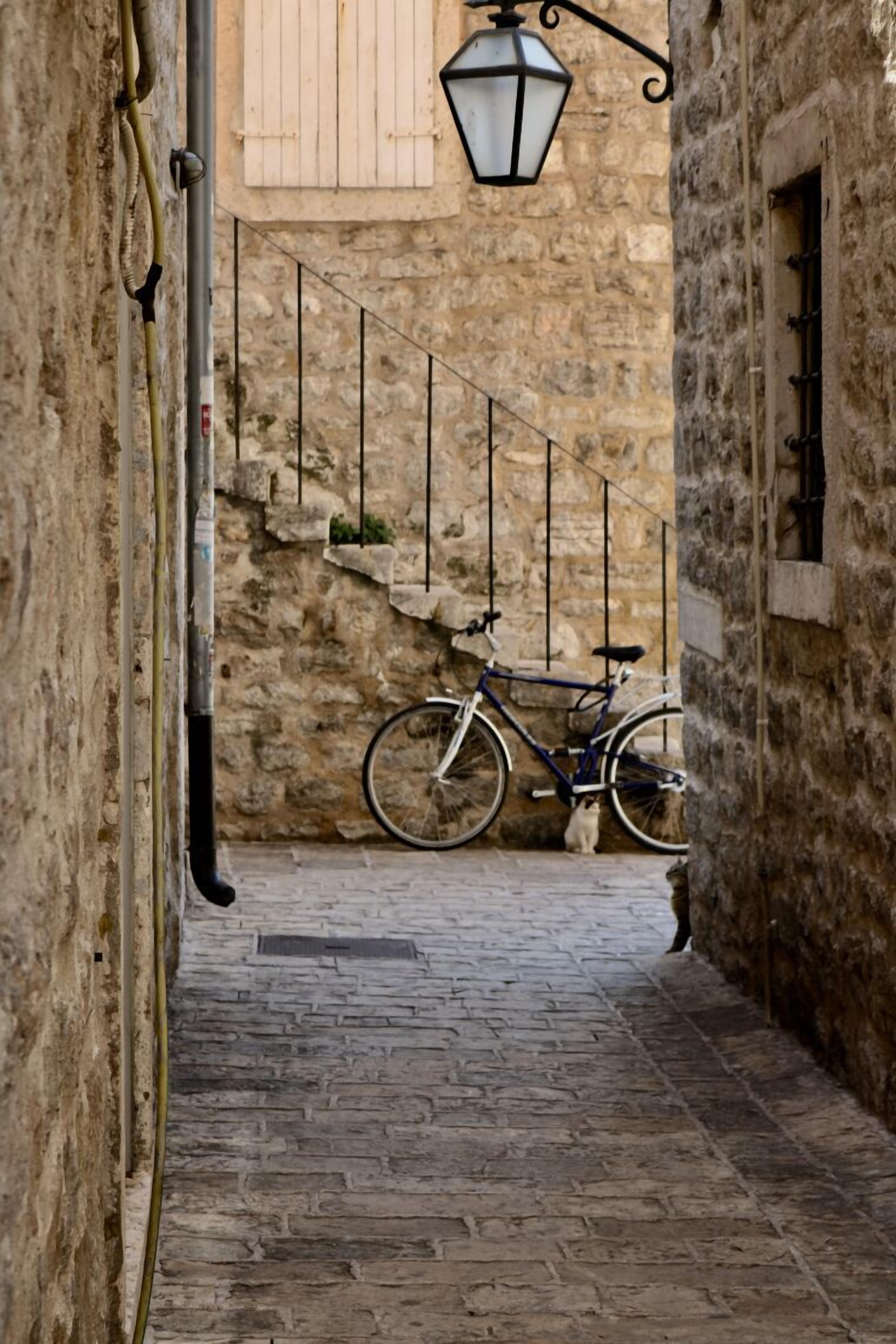 Une ruelle étroite, pavée de pierres, où un vélo est appuyé contre un mur, avec une rambarde en métal et une lanterne suspendue.