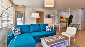 A modern living room with a blue sectional sofa, a white armchair, and a coffee table. There is a kitchen area in the background with white cabinets and a dining table.