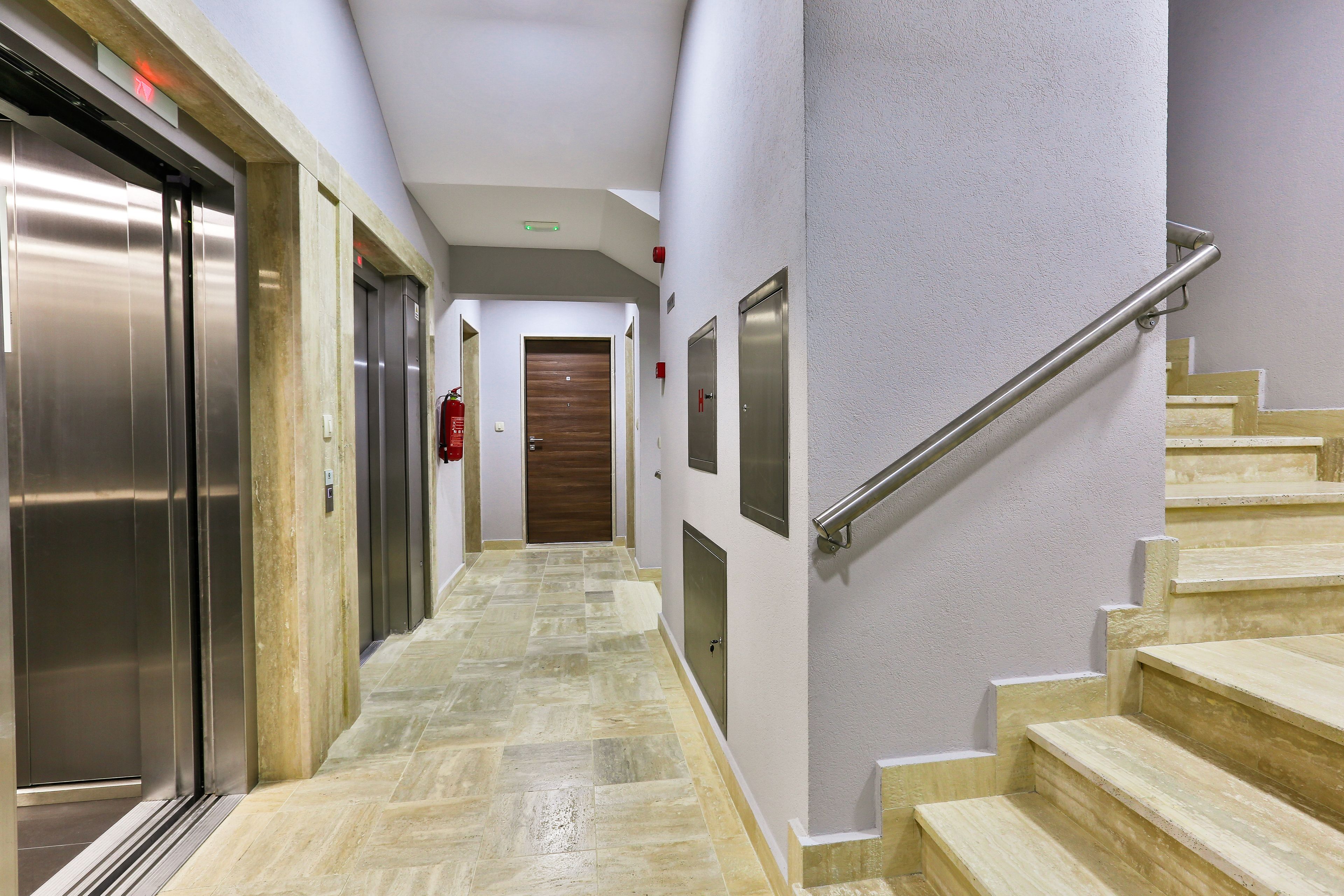 A hallway with elevators, a wooden door at the end, and a staircase with a handrail.