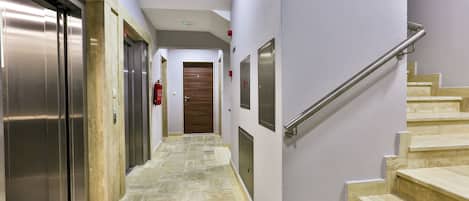 Ein Flur mit Aufzügen, einer Holztür am Ende und einer Treppe mit Handlauf.