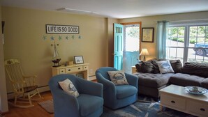 Ein Wohnzimmer mit einem blauen Sofa, zwei blauen Sesseln, einem hölzernen Schaukelstuhl, einem weißen Beistelltisch und einem großen Fenster mit Blick nach draußen.