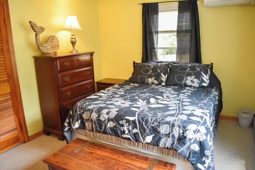 A bedroom with a bed, a dresser, a lamp, a window with curtains, and a small trash bin.