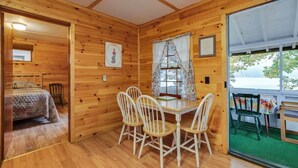 A wooden cabin with a dining area featuring a table and four chairs, a bed with a quilt, and a large window with curtains.