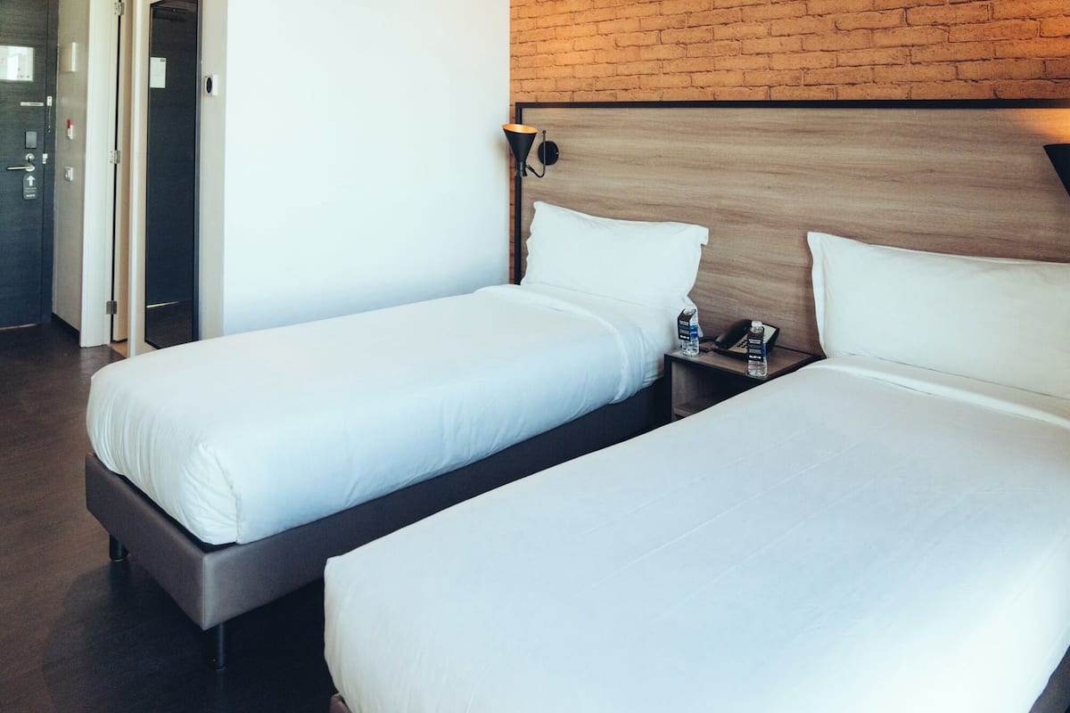 Une chambre d’hôtel avec deux lits simples, une tête de lit en bois, une table de chevet avec un téléphone et une bouteille d’eau, et un mur en briques.