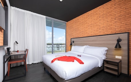 A modern hotel room with a large bed, a desk with a chair, a nightstand, and a view of the outdoors through a large window.