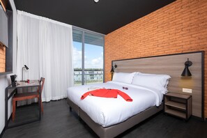 A modern hotel room with a large bed, a desk with a chair, a nightstand, and a view of the outdoors through a large window. - ONOMO Hotel Casablanca Airport (Nouaceur)